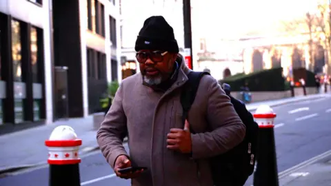 Benjamin Aninakwa pictured walking towards the camera on his way along a busy road into the Old Bailey, wearing glasses, a hat and coat, and carrying a shoulder bag.