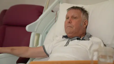 BBC Jabez Petherick, who has short grey hair and is wearing a white polo shirt with grey collar while lying on a hospice bed.