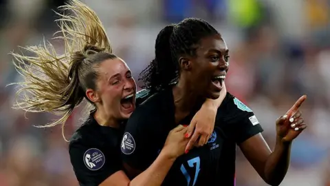 Getty Images Two England players with faces full of joy and wide smiles, both wearing dark blue short sleeve team shirts. With one's blonde pony tile flying wildly. 
