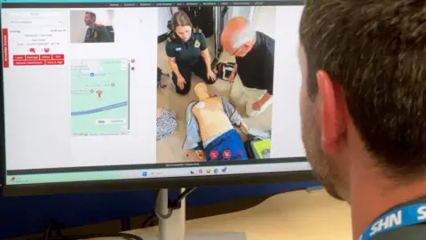 NIKKI FOX/BBC An advanced paramedic looks at his computer screen and sees the moment when ambulance staff arrive to help a bystander who has witnessed someone have a cardiac arrest and has been doing CPR. He is looking at the screen with his head and shoulders and NHS lanyard showing to the right of screen. The computer screen with the image of the dummy patient and bystander is on the left. The computer also shows a map of the location where the patient is and an image of himself.