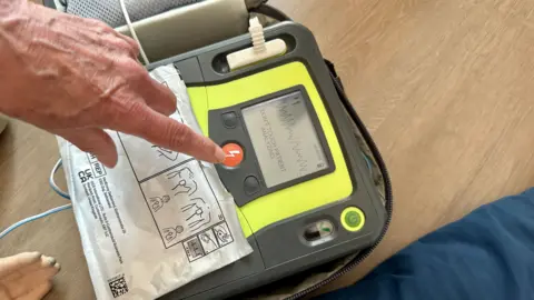 STEVE HUBBARD/BBC A defibrillator on the floor being used. It has a fluorescent yellow and black casing and a finger is floating over the power button. You can't see the person inthe picture. There are instructions on a large white sticker over the front of the device.