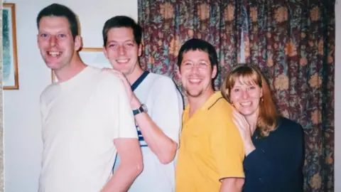 Supplied Tim, Andy, Richard and Sue stand in height order sometime in the 1990s. Tim and Andy are both wearing white shirts while Richard is in a bright yellow shirt and wears glasses. Sue has shoulder length blonde hair and a dark blue top. They are all smiling widely and there is a red and yellow floral patterned curtain in the background.