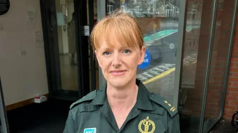 Yvonne McMichael has ginger hair which is tied up with a fringe. She has blue eyes and is wearing pink lipstick and is smiling at the camera. She is wearing a green ambulance service uniform.