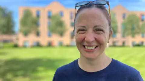 Close up of Kirsty Elliott Sale wearing a blue t-shirt, glasses resting on the top of her brown hair and smiling at the camera against the backdrop of brick buildings and green grass. 