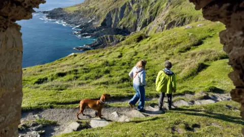 Getty Images Two people are walking along a grassy path accompanied by a brown dog. The path winds toward dramatic, rocky cliffs that drop into the sea.