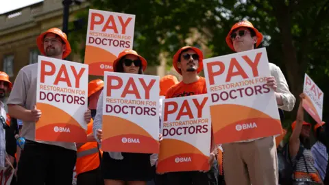 PA Media Three young men and a woman stand in a line at a doctors' strike. They are all wearing identical orange bucket hats and holding placards towards the camera which say 'pay restoration for doctors'. A tree and a crowd of protesters are visible behind them.