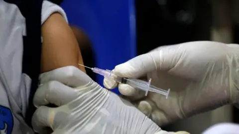 EPA A person wearing white medical gloves injects a needle containing a clear, colourless vaccine into a child's arm. 