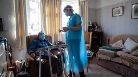 Getty Images A healthcare worker standing and dressed in a blue gown chats to a housebound patient in his front room. The patient is sat down on a chair with a medical walking frame in front of him. He is wearing a mask. There is a sofa on the right of the picture and sun is coming through the windows behind.