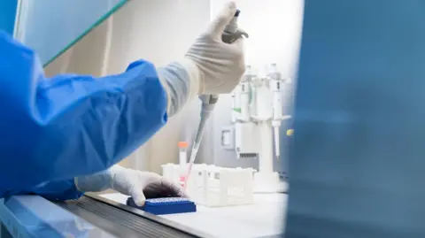 Getty Images Laboratory worker in blue protective clothing and white gloves working with lab equipment and containers on the workbench