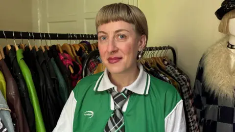BBC A woman with short blonde hair standing in front of rails of jackets and a mannequin