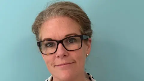 Dr Rebecca Towner Dr Rebecca Towner with brown hair which is tied back, glasses and pearl earrings smiling at the camera