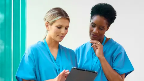 Getty Images A stock photo shows two healthcare professionals in blue scrubs looking at a tablet in a hospital.