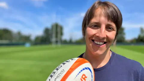 Katy is on the luscious green practice fields with the rugby goal in the background, she stands in the fore-ground, smiling and holding a white an orange rugby ball