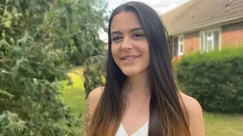 Abbie Abbie standing in a garden in front of a house. She has long brown hair and is wearing a white vest top