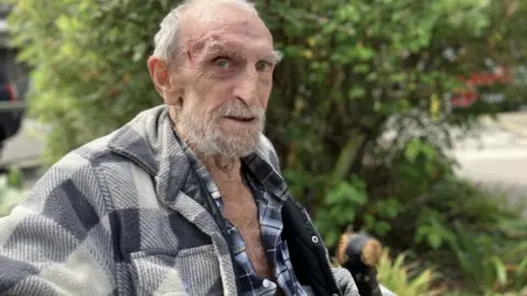 BBC Eddie Baker wearing a checked shirt and jacket. He has white hair, a white beard and is elderly.