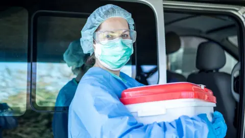 Getty Images a female medic wearing blue scrubs and a face mask holds an organ transplant container, which is a white plastic tub with a red lid. She appears to be stood to the side of a vehicle with a sliding door