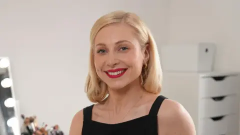 BBC Amy, who has shoulder length blonde hair and wears a sleeveless black top, smiles at the camera. Behind her is a mirror and a number of makeup brushes
