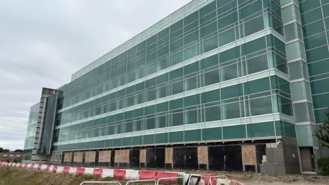 Ben Schofield/BBC Modern multi-story building with glass windows, the ground floor us under construction with boarded-up sections.