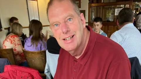 Supplied Tim Hull in what appears to be a pub. He is smiling and wearing a red shirt. His face looks fuller than in other pictures