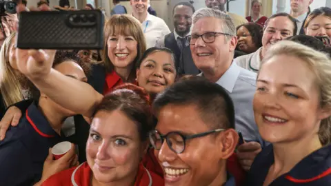PA Media Close up Sir Keir Starmer and Rachel Reeves posing for a selfie taken by an NHS staff member. They are smiling and surrounded by a group of NHS staff all smiling at the camera
