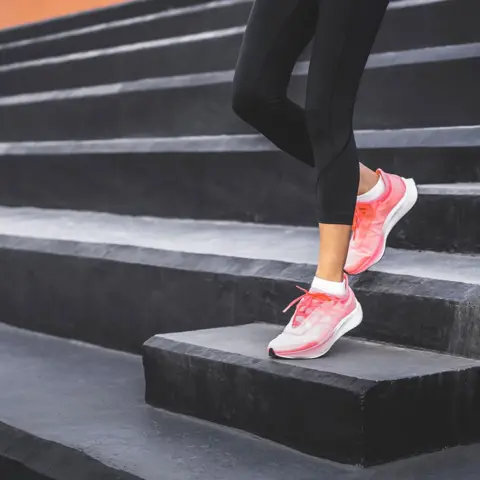 Getty Images A woman wearing black leggings and pink athletic shoes, standing on a set of black steps. One foot is placed on a higher step while the other remains on a lower one, suggesting an exercise or stretching.