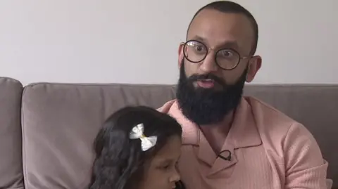 Mr Shahid sits on a grey sofa, wearing a pink shirt and black rimmed glasses. His daughter is sitting with him and wears a white bow in her hair.