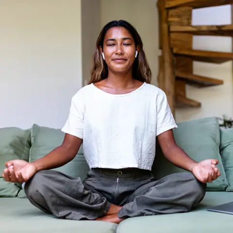 Getty Images A woman sitting cross-legged on a green sofa. She is wearing a white T-shirt, grey trousers, and wireless earbuds. In the background, there is a wooden spiral staircase.
