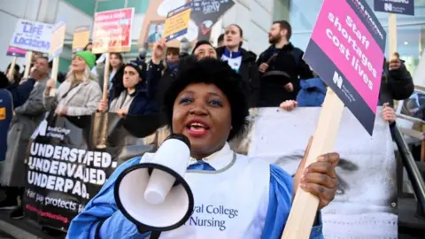 Reuters A nurse, holding a megaphone in one hand and a Royal College of Nursing placard in the other, stands on a picket line outside UCH hospital in London in 2023. She is standing in front of other pickets who are holding brightly coloured banners. 