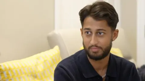 24-year-old Amrit sat on his sofa with a straight face as he is interviewed for BBC London. He has a groomed beard and wears a navy shirt.