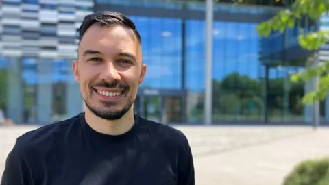 Young man wearing a dark T-shirt stands in front of a shiny glass building on a hot sunny day, smiling, short hair and closely cropped beard and moustache. 