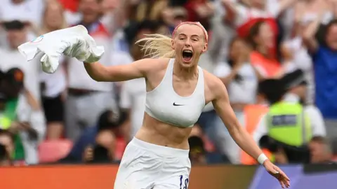 Getty Images Chloe running towards the camera with her arms to her sides, wearing white shorts and white sports bra with her England shirt twirled up in her right hand, screaming with joy, blonde hair flying