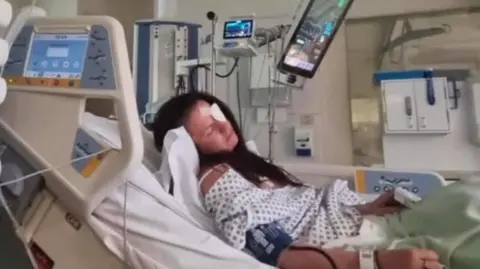 Family handout Kaylie lying on a hospital bed surrounded by machines. She has long brown hair and a white patch over her left eye. She is wearing a white gown and her head is at an angle suggesting she is asleep.