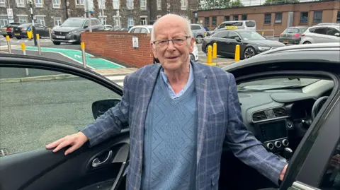 Tommy Nicholl is an elderly man who has white balding hair. He has dark glasses on and is wearing a blue shirt, jumper and blazer. He is standing in a car park with one arm inside a dark green car and the other arm resting on the door which is open.