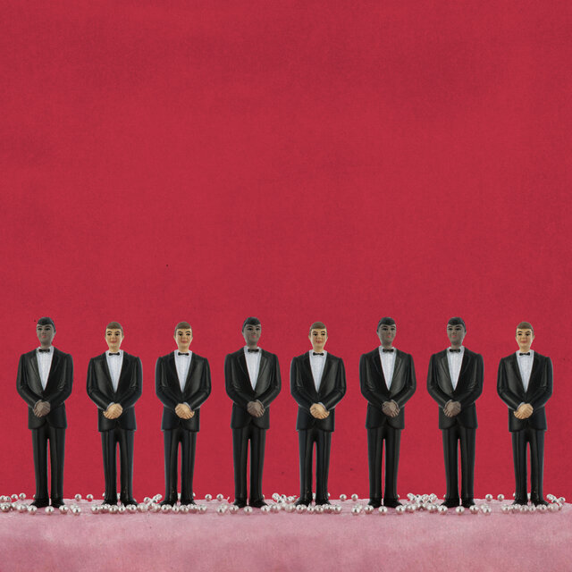 Eight figurines of men in tuxedos standing in a line on a red background. 