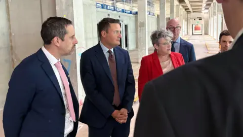 Ben Schofield/BBC Wes Streeting and a group of people in formal attire are standing in an unfinished building, with concrete wall in the background and visible construction materials