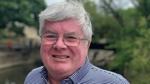 Willie is smiling outside near a river bank with trees. He has thick, short white/grey hair and is wearing thin framed glasses. He has a dark blue and white striped shirt on. 