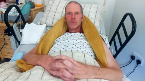 BBC Tim Hull lies in bed with a yellow blanket round his neck and shoulders. He has blue eyes and short hair with his hands folded across his lap