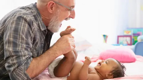 Getty Images An older dad with a grey beard and wearing glasses and a checked shirt, holds a young baby's feet while she lies on a bed, wearing a nappy, and smiles up at him
