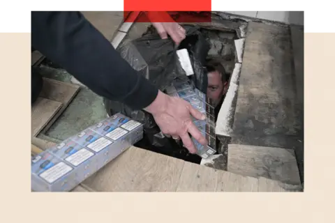 BBC/Phillip Edwards An officer bring out illegal packets of cigarettes from a hole in the floor following a raid on a corner shop