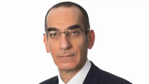 BBC A man who is about 50, with short dark shaved hair, distinctive dark eyebrows and rimless glasses. He is looking straight at the camera and is not smiling. He is pictured - head and shoulders - against a plain white background. 
