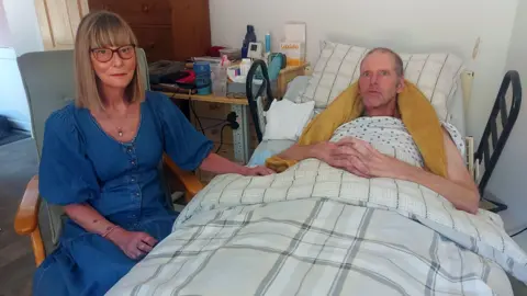 Sue and Tim Hull. Sue has shoulder-length straight blonde hair and is wearing a blue dress and glasses. She is sitting next to Tim who is lying in bed. He has his arms folded across his lap and a yellow blanket round his neck and shoulders.