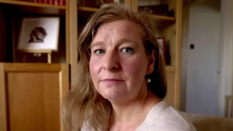 BBC A headshot of Sarah, looking straight at the camera with a serious expression. She is wearing an oat coloured top and earrings with her hair loose. She is sitting in a living room, there are wooden shelves behind her. 