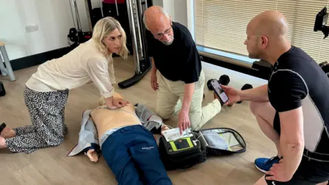 STEVE HUBBARD/BBC A wide picture showing two bystanders and a community first responder gathered around a dummy. A woman with blonde hair kneels down over the dummy doing CPR while she looks at a man on a video call to the east of england ambulance service advanced paramedic. A first responder has a defibrillator on the floor and they are attempting to use it. A wire is attached from the defibrillator to the machine. They are all on their knees in a gym.