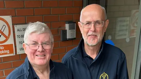 BBC Willie Hutchinson is in his 70s and has grey hair and glasses. He is standing beside Martin Garrity who is bald with a grey beard and glasses. They are both wearing navy jackets and polo shirts and are smiling at the camera.