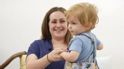 University Hospital Southampton Ceri is wearing a dark blue top and is smiling at her son Harry, while holding his hands. He is wearing a light blue T-shirt and colourful dungarees and has wavy, auburn hair, and is turning to the side.