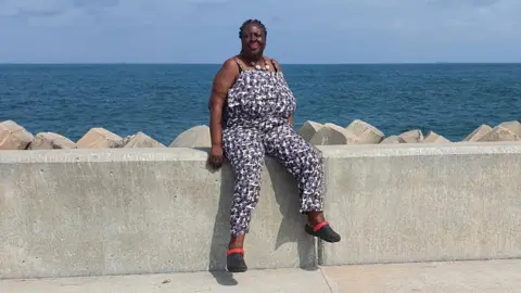 Supplied A woman in a pattern jumpsuit and sandals sits on a wall infront of the ocean, posing and smiling for the picture
