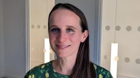 BBC Dr Emily Jones, who has with long brown hair and blue eyes, is smiling. She is wearing a green floral top with yellow and green patterns and is looking into the camera. The background is a mixture of white walls and glass with a dotted design and lights hanging from the ceiling behind it. 