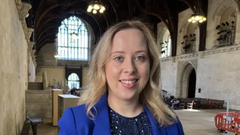 Becky Gittins, who has wavy blonde hair and is wearing a blue suit jacket with a blue animal print dress, smiles at the camera