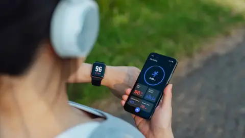 Getty Images A person is holding a smartphone displaying a fitness tracking app while wearing white headphones and a smartwatch.