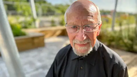Nikki Fox/BBC John Newman looks at the camera. His head and shoulders are visible. He is wearing a black shirt and has glasses on. He has blue eyes and a tanned complexion with a bald head and some grey hair to the side with a grey moustache and beard.